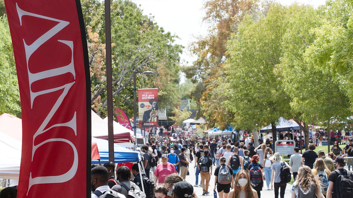 Students walking on campus with booths and tents aligned along the walkway while a red UNLV banner is in the foreground