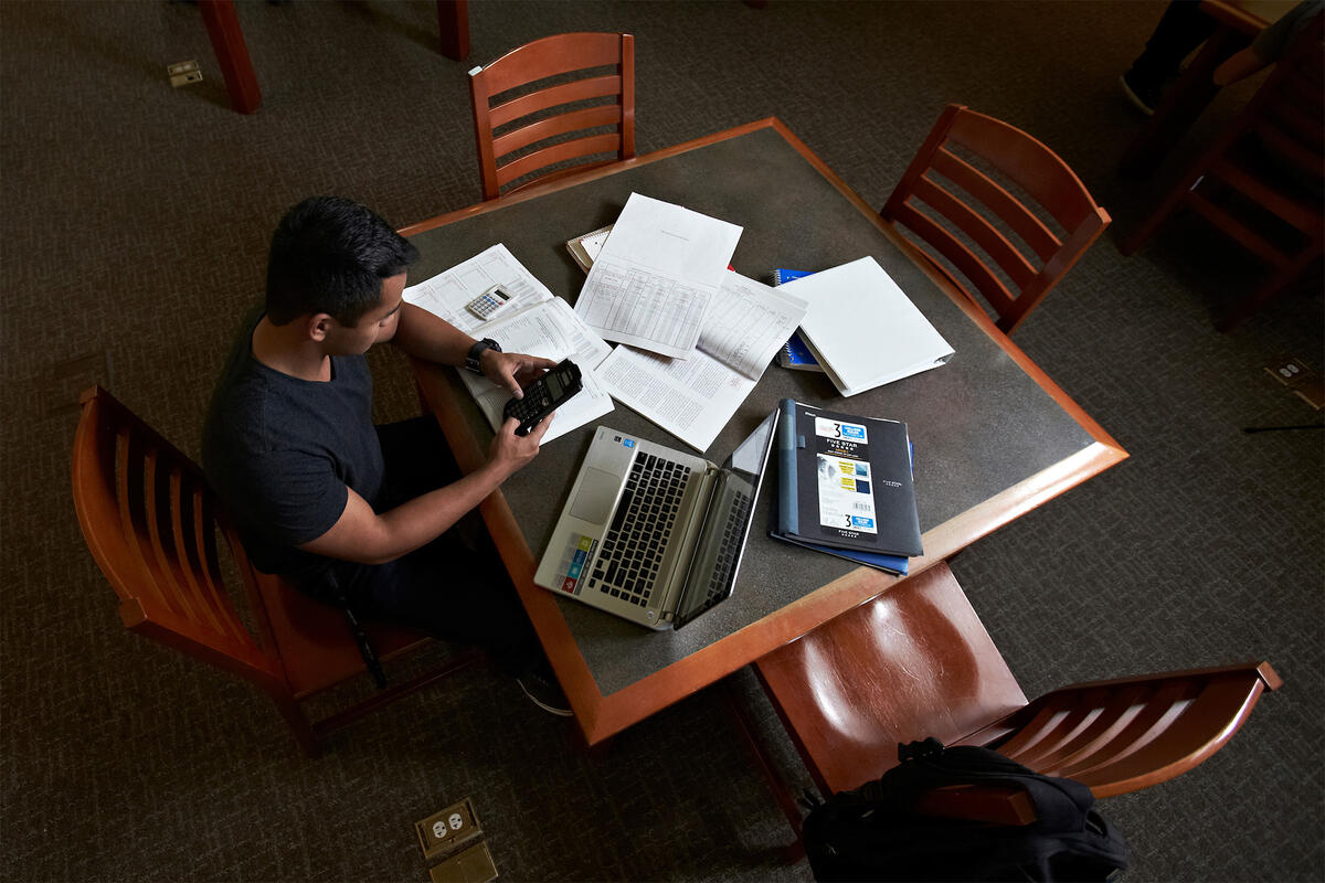 A person at a table studying with a laptop and a pile of paper scattered on it