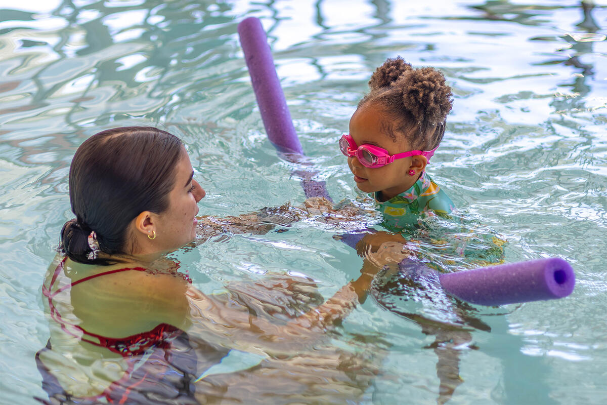 A student lifeguard helping a child with a purple pool noodle swim across the pool