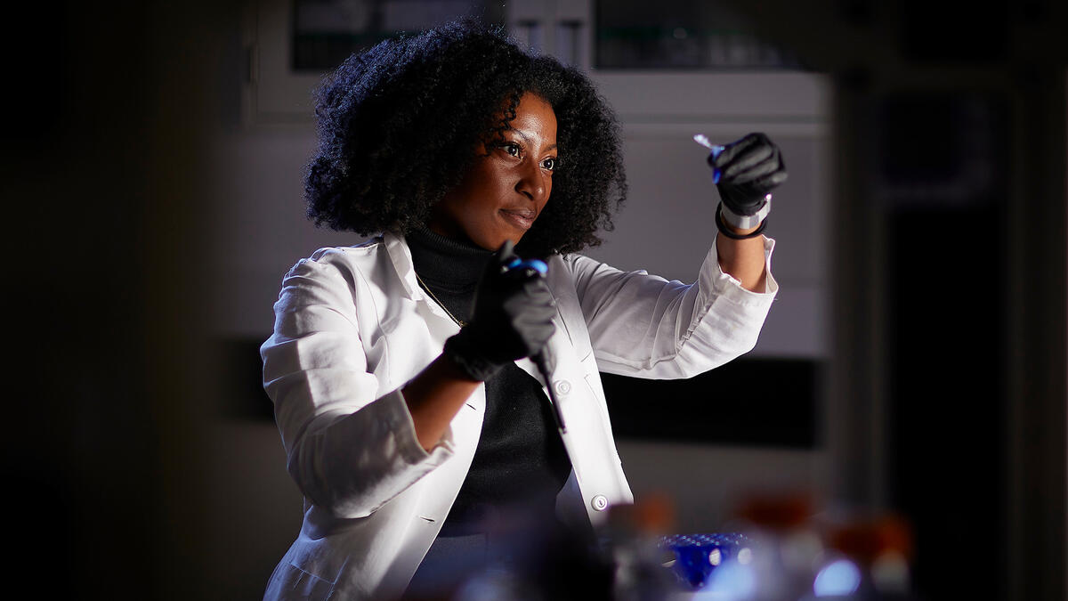 A woman wearing a white lab coat and protective goggles holding up a lab sample