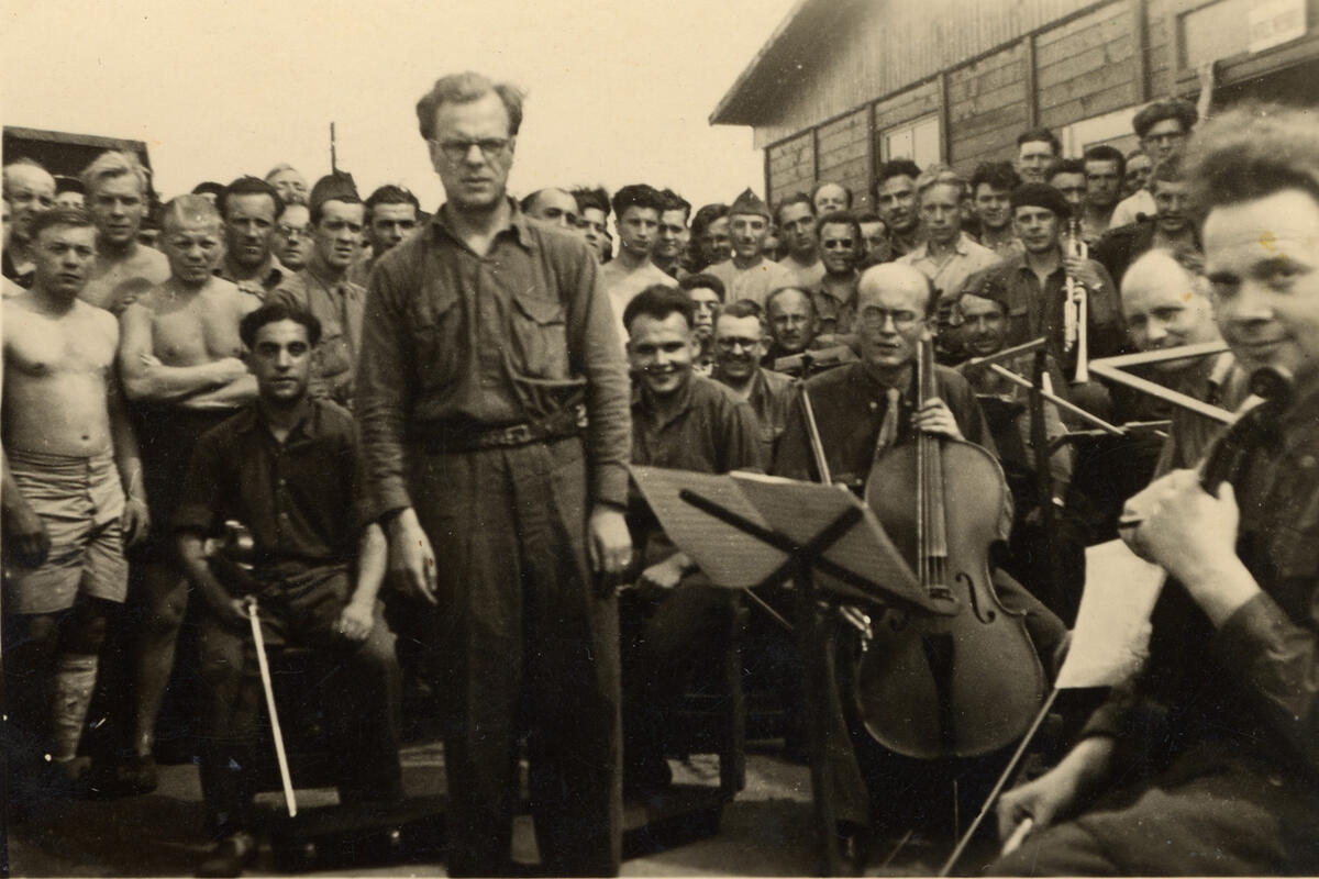 The image is a picture of a conductor, holding his baton, surrounded by instrumentalists. They appear to be in a camp for prisoners of war.