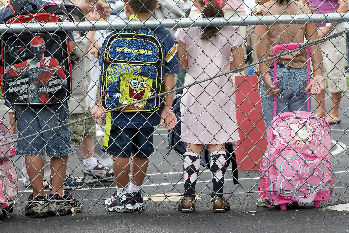 children lean against chain-linked fense with backpacks