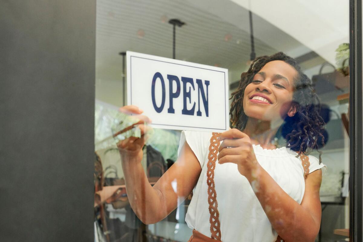 smiling black woman hangs "open" sign on the window of her business