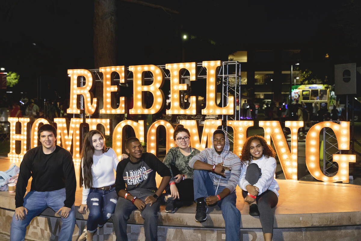 students pose in front of lighted sign that reads Rebel Homecoming