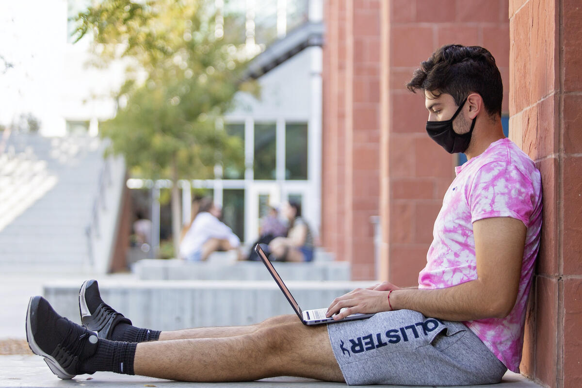 student wearing face covering sits on ground, using laptop