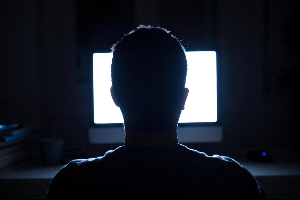 Silhouette of man sitting in a dark room in front of a glowing computer screen.