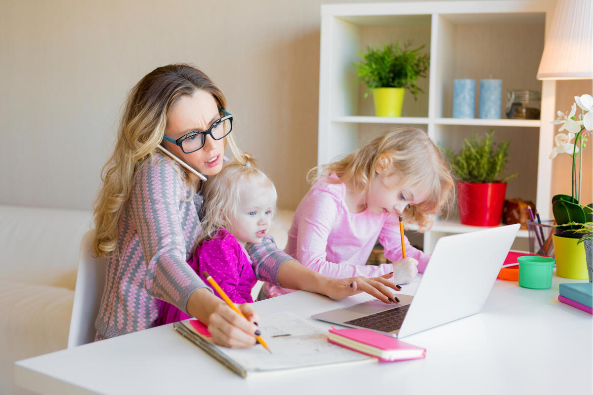 stressed woman works on computer with toddler on lap and slightly older child sitting next to her doing homework