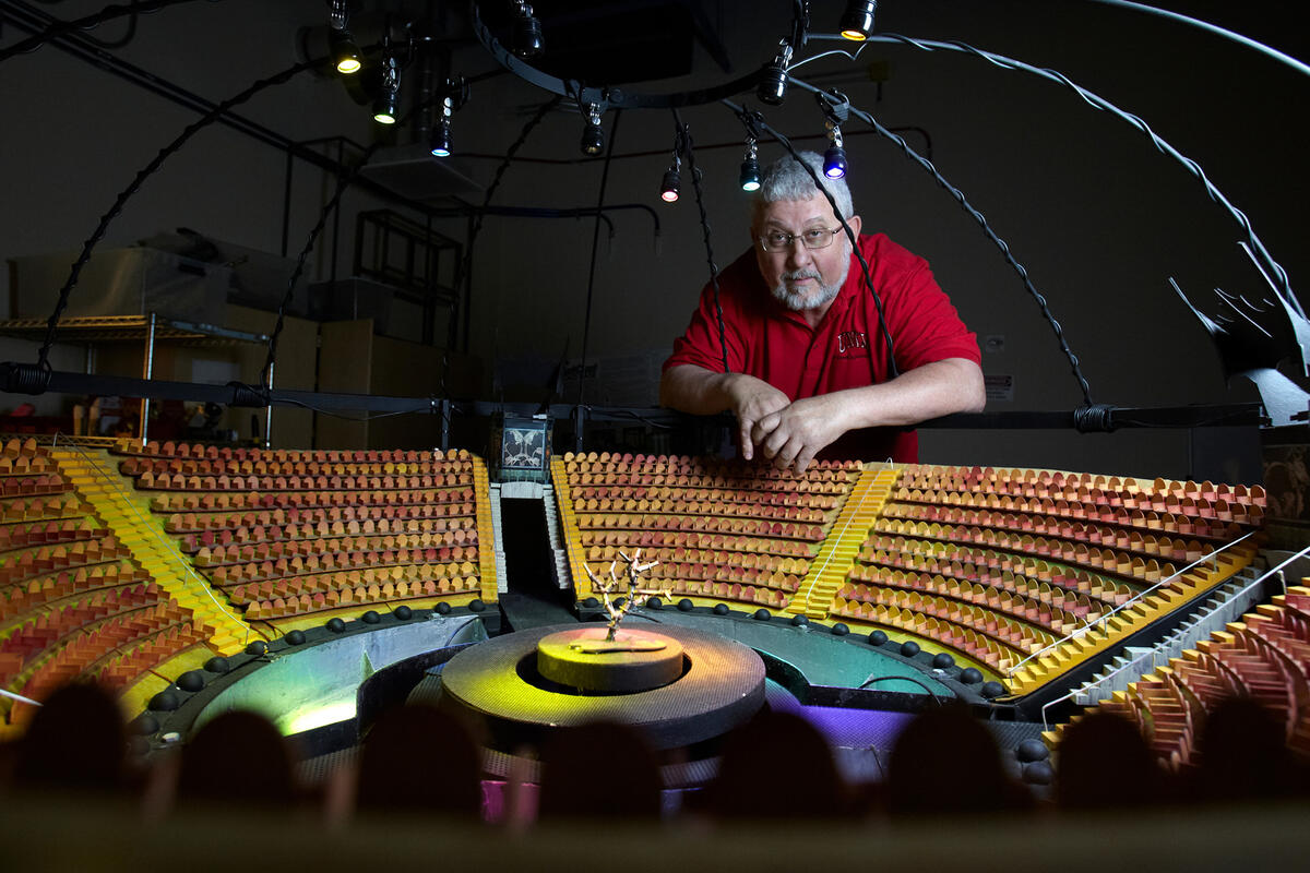 A man working on a miniature scale version of an entertainment venue auditorium with seats encircling a stage and L.E.D. lights shining towards the center.