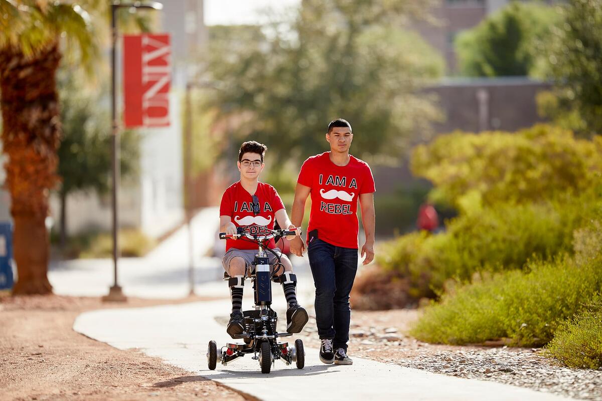 UNLV students going through campus one student rides a scooter while the other walks