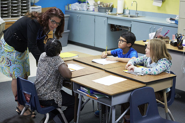 Female teacher bending over to talk to three elementary-aged children sitting at their desks