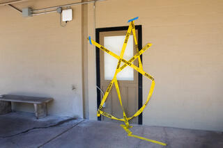 Festoons of yellow caution tape have been fastened haphazardly over a wall panel. The panel is the same size and shape as a door, but it has no handle or hinges. The top half of the panel is a blank white window. A dripping tap and a cracked bench stand nearby on grubby concrete.