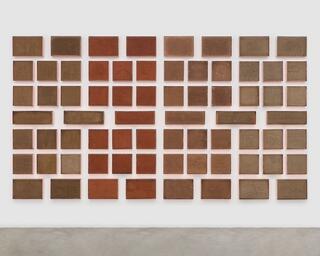 A mass of earth-brown squares and rectangles is arranged in seven rows on a wall. They form a block of modulated hues—the ones on the right are a lighter shade of brown, the one on the left are darker, and the ones in between become reddish—with a grid of white gaps running between their edges. Line drawings of animals, landscapes, and urban infrastructure have been incised into the brown surfaces. Some are cracked like dry earth.
