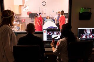 Three clinicians observe diagnostic screens and students in a simulated hospital room from behind glass. On the other side of the observation glass, students tend to a manikin.