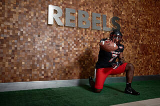 An athlete in Rebel football gear holds out a football in front of a wall with the word, &quot;Rebel&quot;