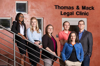 Six students pose along a staircase in front of a "Thomas &amp; Mack Legal Clinic" sign.