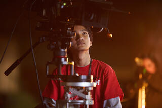 A UNLV Film student in a red shirt directs from behind a film camera.