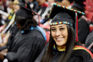 A graduate student wears a black graduation cap and gown, with tribal beading adorning the cap.