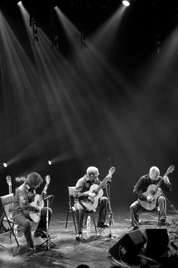 Black and white photo of Montreal Guitar Trio performing. Three men on stage, sitting, while playing guitars.
