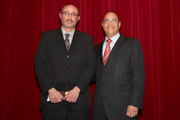 Two men stand in front of a red curtain and smile for the camera.