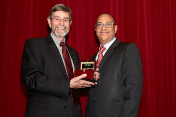 Two men standing in front of a red curtain both smiling while one holds an award.