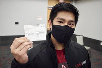 Paul poses with a facemask and holds a vaccine card up to the camera