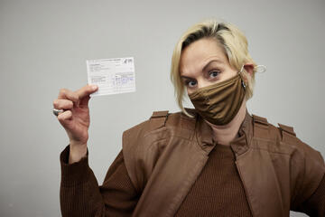 A person in a mask poses with one eybrow raised and holds their vaccine card up to the camera