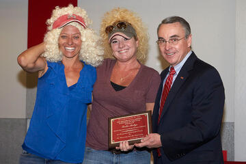 Three people posing for picture. Two are wearing wigs. Person in center is holding an accepted award plaque.