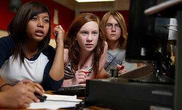 Three young students staring intensely at a single computer monitor.