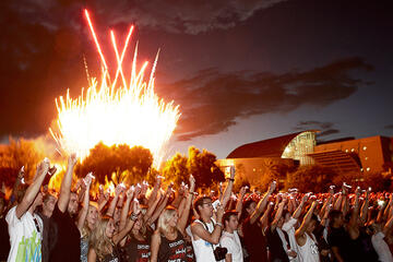 Fireworks exploding behind a crowd of students all raising their hands.