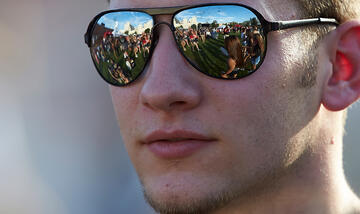 Close up view of guy with sunglasses on. A reflection of the crowd of people that he is looking at is seen in his sunglasses.