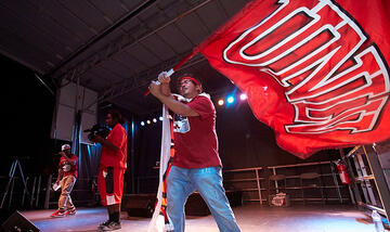 Front side view of a performance on stage. One man is waving a UNLV flag. Two others are singing inside their microphones.