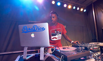 Front side view of a dj as he focuses on his laptop with his right hand and controls a mixer with his left.