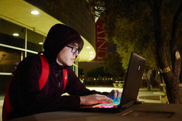 A student works on a laptop on a picnic table in front of a library at night.