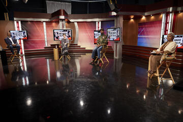 A number of panelists sit in chairs separated by several feet while being filmed.