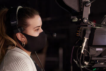 A student wears headphones and a mask, looking through a large film camera