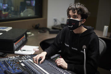 A student operates a sound board while wearing a mask with an embroidered cat face.