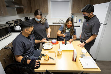 Four UNLV occupational therapy doctoral students wearing dark gray scrubs work around a kitchen table with adaptive knives and other tools