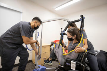Two students wearing gray scrubs use a Hoyer lift to place a third student in a wheelchair during a class demonstration
