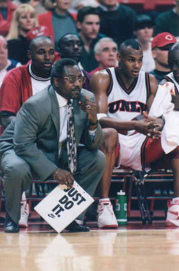 A man in a suit leans forward from the bench of a basketball court