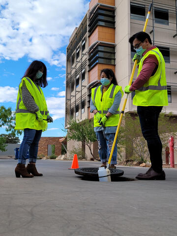 Three UNLV researchers in construction vests and gloves stand around a manhole cover to simulate sewage collection