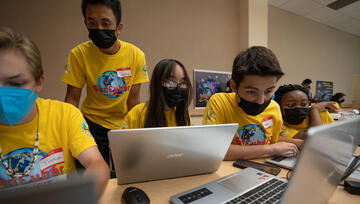 A group of five high schools in yellow t-shirts hover around several laptops to participate in cybersecurity activities
