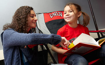 Student pointing to a word inside of a book that a young child is holding.