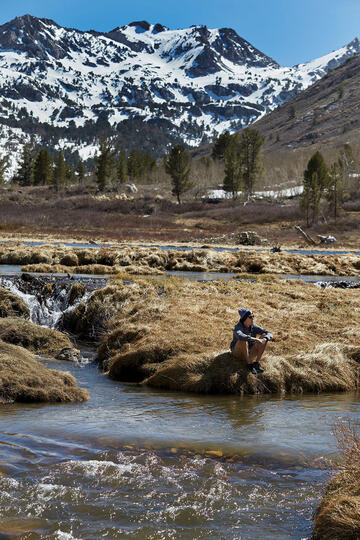 Man near river with snow mountains behind.