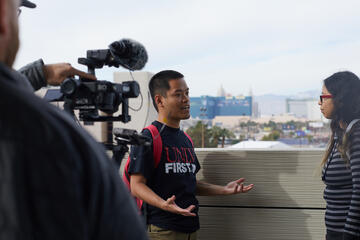 On a rooftop patio, Hieu Nguyen speaks to another student while being filmed with a video camera.