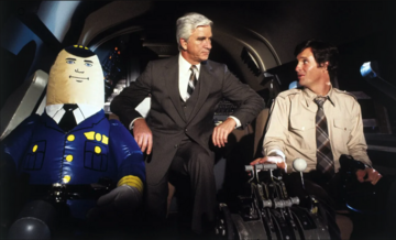 Screenshot from the 1980 movie Airplane! An inflatable doll in a pilot costume, Leslie Nielsen, and Robert Hays sit in a plane's cockpit.