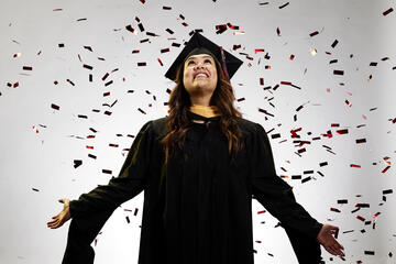 Woman in a black graduation cap and gown with confetti falling