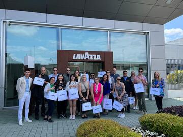 A group of students stand in front of the Lavazza factory. They are all holding Lavazza gift bags.