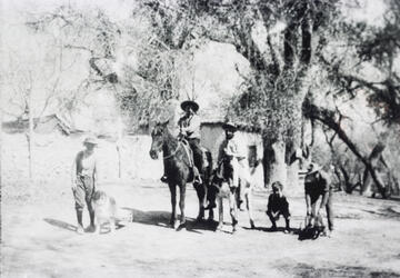 1909 black and white image of men and a boy, horses and dogs outside of buildings at the Mormon Fort, Las Vegas, Nevada.