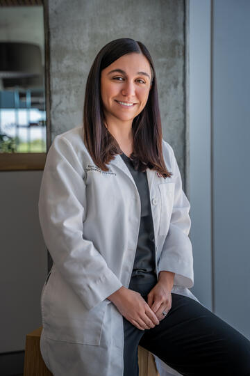 White woman with straight brown hair and wearing a white medical coat smiles as she poses with her left leg slightly bent and her hands crossed below her waist