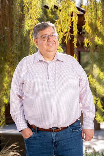 White man in light-colored plaid button-down shirt and blue jeans smiles while standing and posing in an outdoor setting with trees behind him